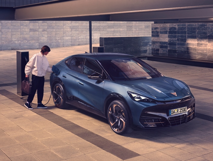 Eine Person lädt einen CUPRA Tavascan, ein vollelektrisches Auto, an einer öffentlich zugänglichen E-Ladestation in einer modernen urbanen Umgebung auf. Das sportliche Elektroauto steht auf einem hell gepflasterten Platz, im Hintergrund moderne Gebäude