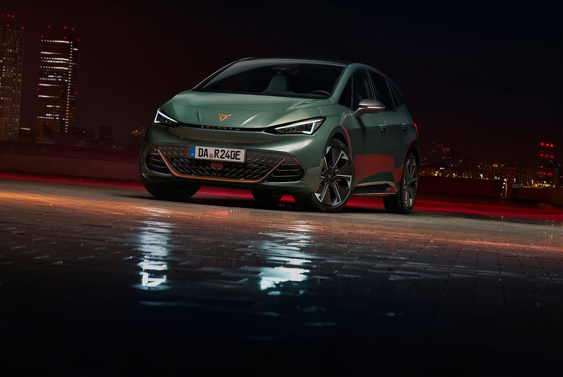 CUPRA Born VZ in mattem Grün, frontal aufgenommen bei Nacht vor einer beleuchteten Stadt-Skyline. Reflektionen auf dem nassen Asphalt betonen das sportliche Design