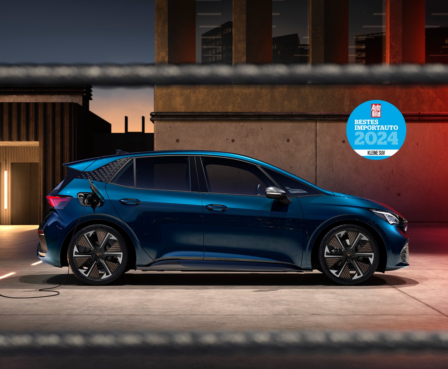 CUPRA Born in seitlicher Ansicht an einer Ladestation in urbaner Umgebung bei Dämmerung. Im Hintergrund ein modernes Gebäude mit warm beleuchteten Türen. Oben rechts eine blaue Auto Bild-Auszeichnung für das beste Importauto 2024